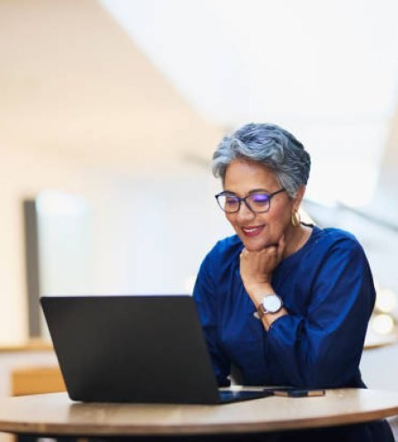 A mature woman with short gray hair and glasses smiles while working on a laptop at a round wooden table. She rests her chin on her hand in a bright, modern office setting.